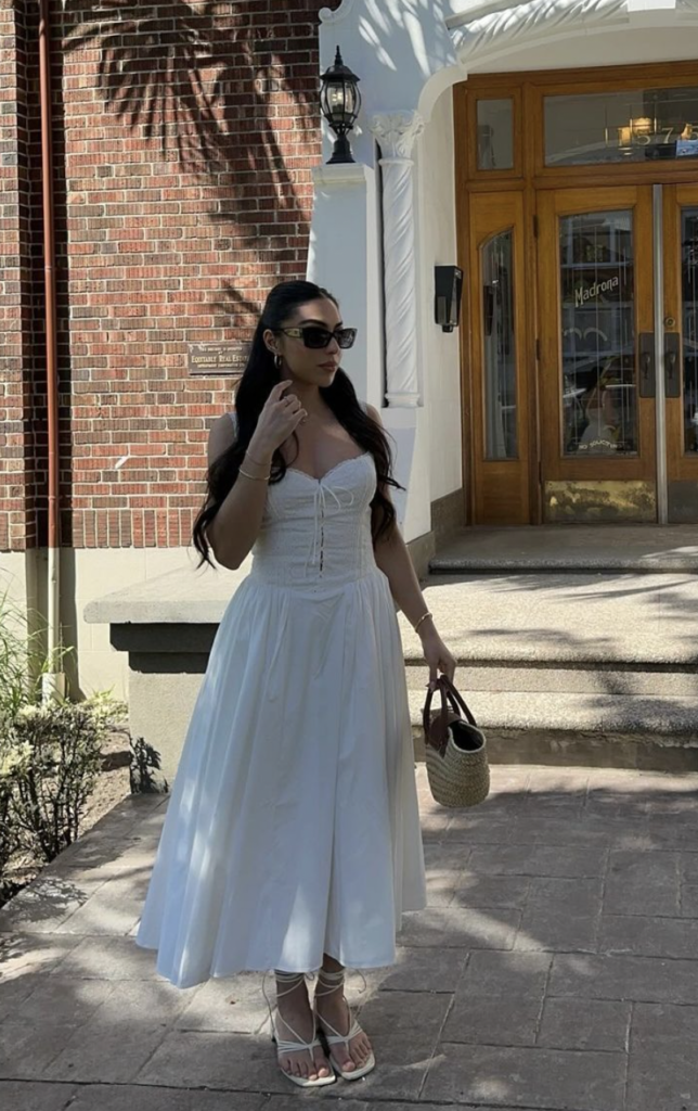 Woman in a white summer dress with lace-up bodice standing in front of a brick building and holding a woven handbag.